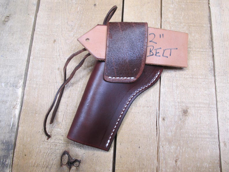 May include: A brown leather holster with white stitching and a belt loop. The holster is designed to fit a 2-inch belt. The image shows the holster on a wooden surface.