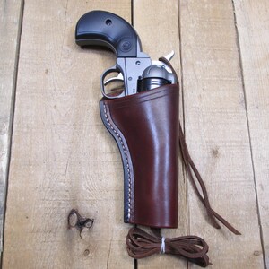 May include: A brown leather holster with a silver revolver inside. The holster has a strap and a loop for attaching to a belt.