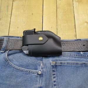 Könnte beinhalten: Ein schwarzes Lederholster mit einem Messingknopf, getragen an einer blauen Jeans mit einem braunen Ledergürtel.