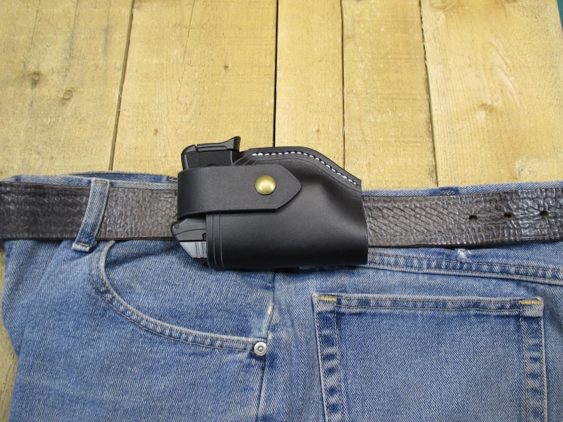 May include: A black leather holster with white stitching and a brass button is attached to a brown leather belt. The belt is worn through the loops of blue denim jeans. The background is a light brown wooden surface.