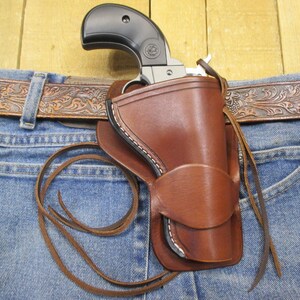 Puede incluir: Una funda de cuero marrón con un revólver negro, sujeta a unos vaqueros azules. La funda tiene costuras blancas y una correa de cuero. Se ve un cinturón de cuero marrón repujado por encima de los vaqueros.