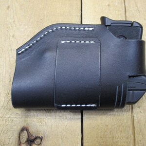 May include: Black leather holster with white stitching. The holster is designed to hold a small handgun and a magazine. The stitching is visible along the edges and around the belt loop. The holster is shown on a wooden surface.