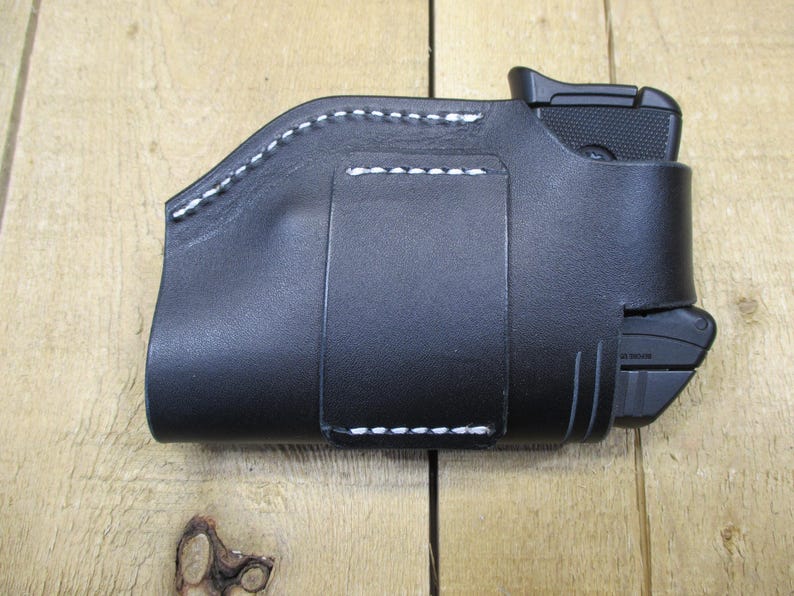 May include: Black leather holster with white stitching. The holster is designed to hold a small handgun and a magazine. The stitching is visible along the edges and around the belt loop. The holster is shown on a wooden surface.