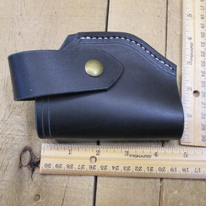 May include: Black leather holster with a strap and brass button closure. The holster is approximately 5 inches long. White stitching accents the top edge. The item is shown on a wooden surface.