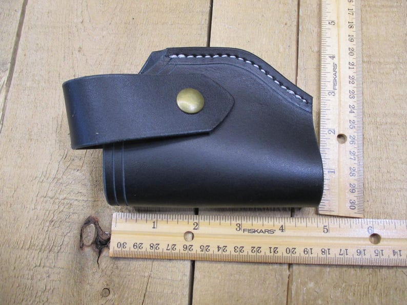 May include: Black leather holster with a strap and brass button closure. The holster is approximately 5 inches long. White stitching accents the top edge. The item is shown on a wooden surface.