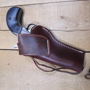 May include: A brown leather holster with white stitching holds a black and silver handgun. The holster has a flap and a leather tie. The background is a light brown wooden surface.