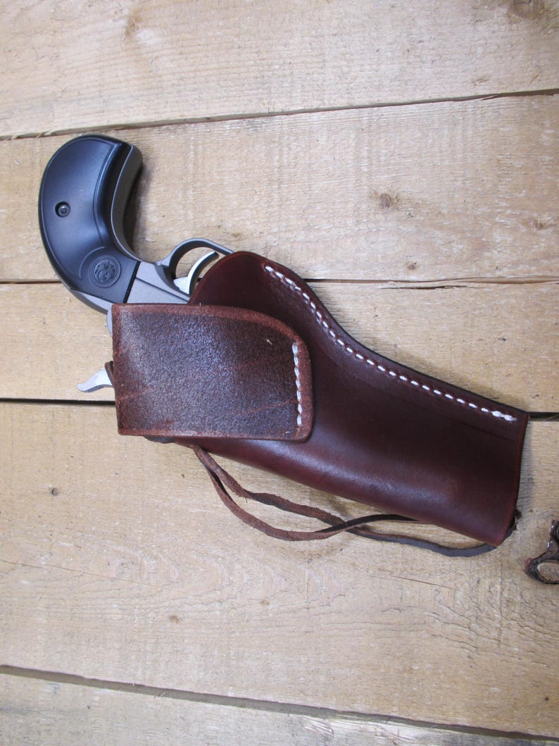 May include: A brown leather holster with white stitching holds a black and silver handgun. The holster has a flap and a leather tie. The background is a light brown wooden surface.