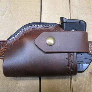 May include: A brown leather holster with a black handgun inside, secured by a strap with a brass button. The holster is attached to a brown leather belt with a floral design. The holster has white stitching.