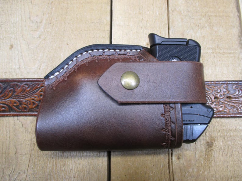 May include: A brown leather holster with a black handgun inside, secured by a strap with a brass button. The holster is attached to a brown leather belt with a floral design. The holster has white stitching.