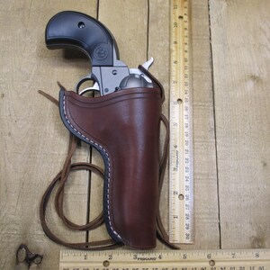 May include: A black and silver revolver in a brown leather holster with white stitching. The holster has a brown leather strap. A wooden ruler is next to the holster, measuring in inches.