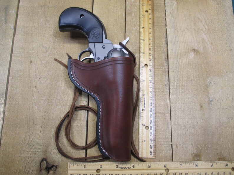 May include: A black and silver revolver in a brown leather holster with white stitching. The holster has a brown leather strap. A wooden ruler is next to the holster, measuring in inches.