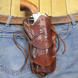 May include: A brown leather holster with a detailed embossed design, holding a silver and wood revolver. The holster is attached to a brown leather belt with an embossed floral pattern, resting on blue denim jeans.