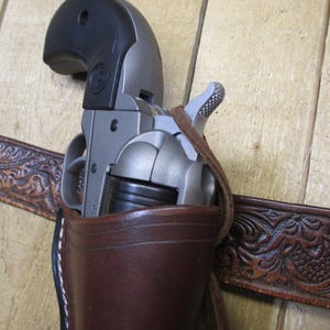 May include: A brown leather holster with a silver and black revolver inside. The holster is attached to a brown leather belt with an embossed floral design.