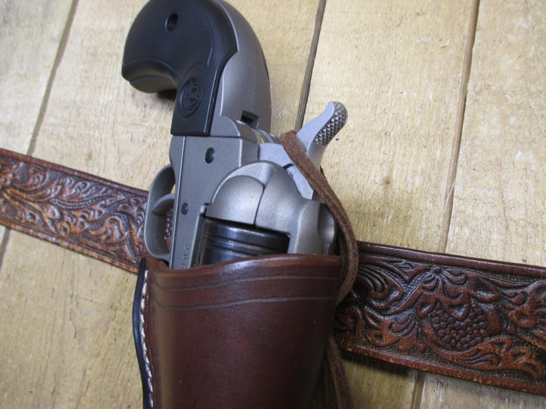 May include: A brown leather holster with a silver and black revolver inside. The holster is attached to a brown leather belt with an embossed floral design.