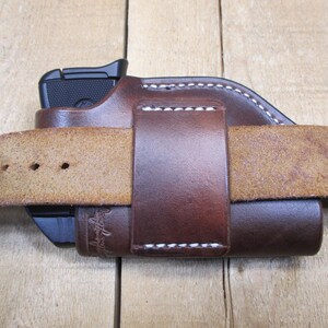 May include: A brown leather holster with a black handgun inside, secured by a matching belt loop. The holster is stitched with white thread and is attached to a light brown leather belt. The image is set against a wooden background.