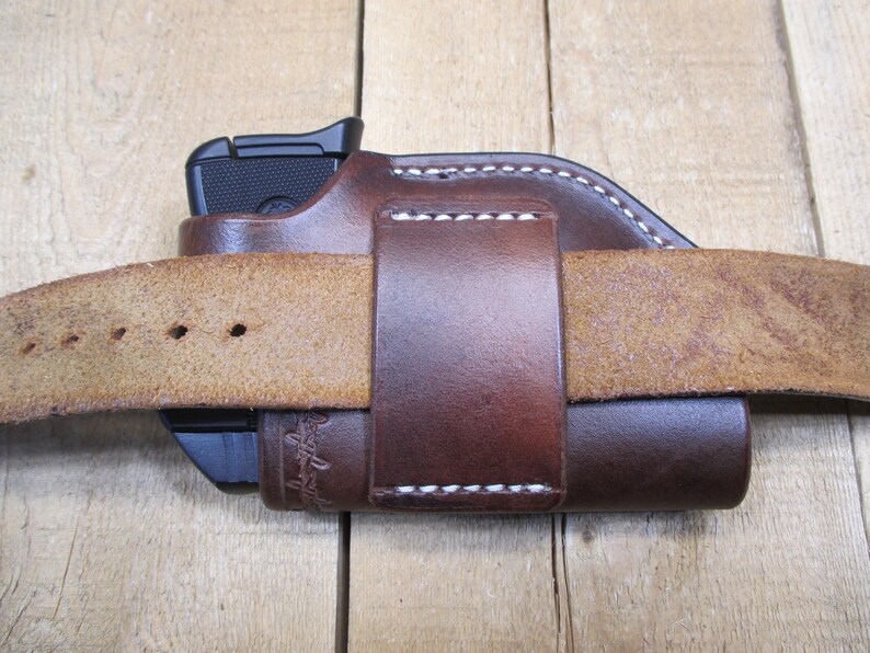 May include: A brown leather holster with a black handgun inside, secured by a matching belt loop. The holster is stitched with white thread and is attached to a light brown leather belt. The image is set against a wooden background.