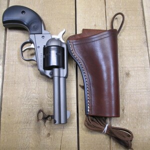 May include: A silver revolver with a black handle and a brown leather holster. The holster has a strap and a loop for attaching to a belt.