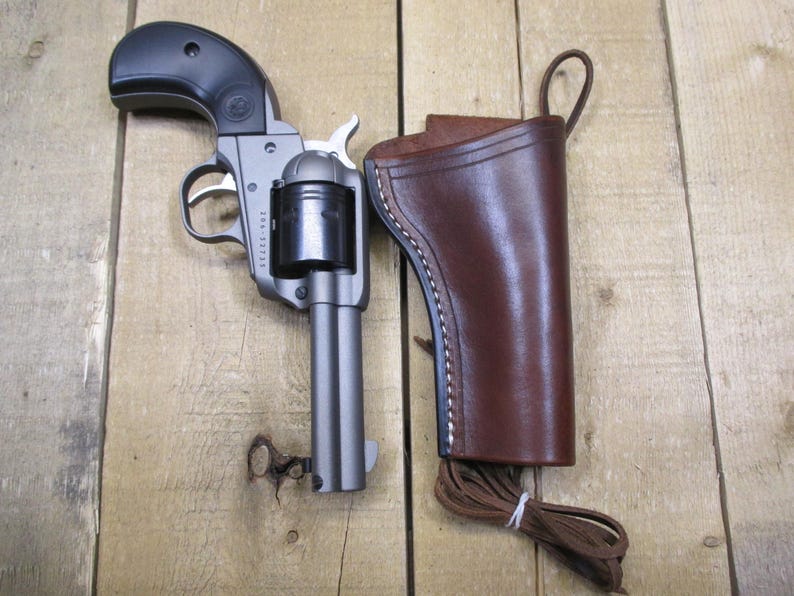 May include: A silver revolver with a black handle and a brown leather holster. The holster has a strap and a loop for attaching to a belt.