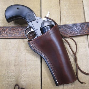 May include: A silver revolver with a black grip is holstered in a brown leather holster. The holster is attached to a brown, tooled leather belt. The background is a wooden surface.