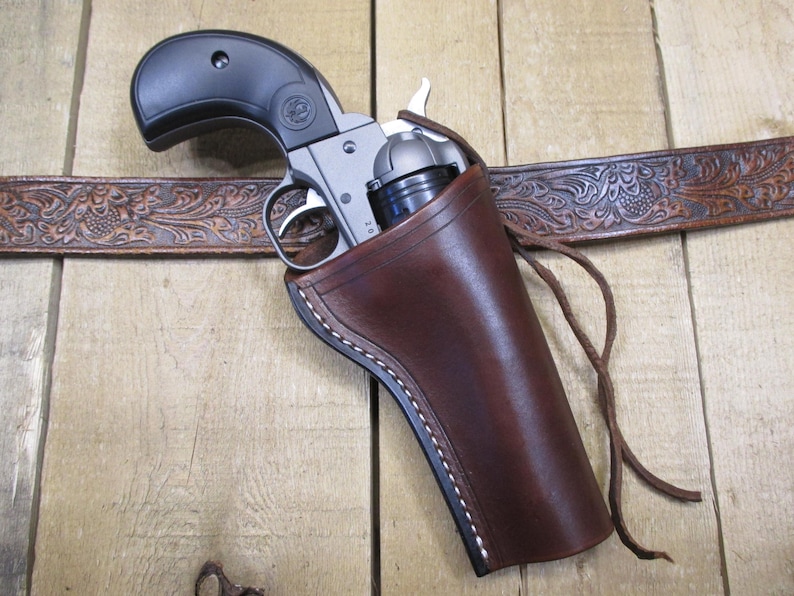 May include: A silver revolver with a black grip is holstered in a brown leather holster. The holster is attached to a brown, tooled leather belt. The background is a wooden surface.