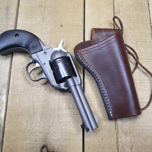 May include: A revolver with a black grip and a silver barrel, next to a brown leather holster with white stitching. The gun is resting on a wooden surface.