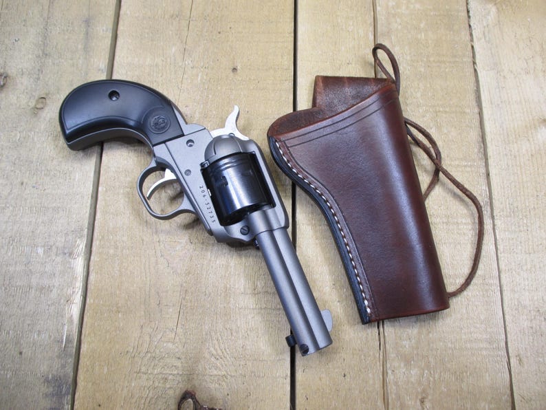 May include: A revolver with a black grip and a silver barrel, next to a brown leather holster with white stitching. The gun is resting on a wooden surface.