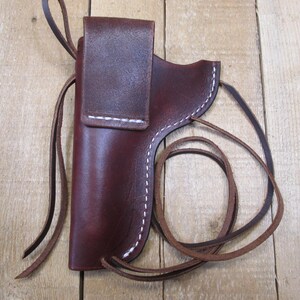 May include: A burgundy leather holster with white stitching and a flap closure. The holster has a loop for a belt and a long leather strap. The item is displayed on a wooden surface.