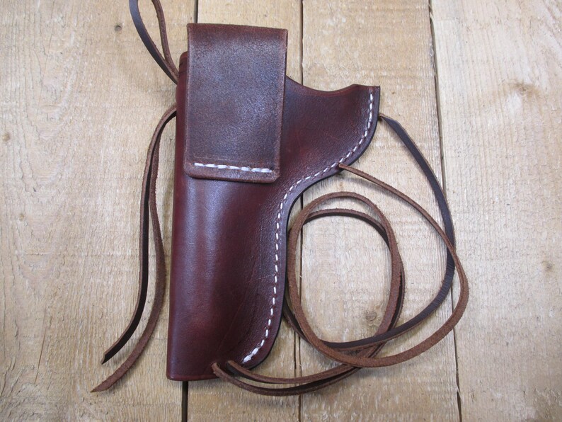 May include: A burgundy leather holster with white stitching and a flap closure. The holster has a loop for a belt and a long leather strap. The item is displayed on a wooden surface.