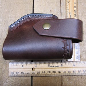 May include: A brown leather holster with a stitched top edge and a flap secured by a brass button. The holster has a curved design and measures approximately 6 inches in length.