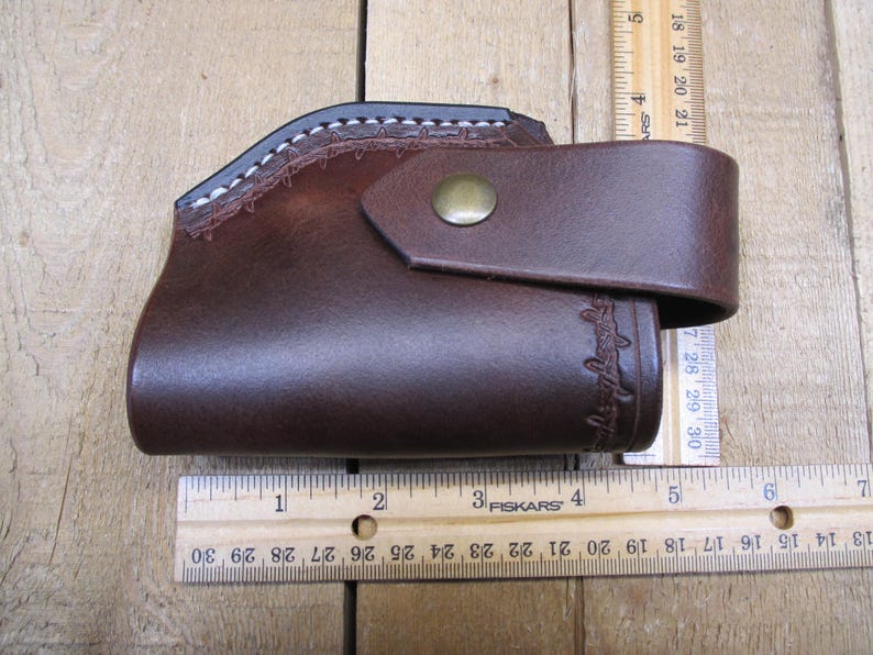 May include: A brown leather holster with a stitched top edge and a flap secured by a brass button. The holster has a curved design and measures approximately 6 inches in length.