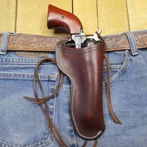 Könnte beinhalten: Ein braunes Lederholster mit einem genähten Rand, das für einen Revolver ausgelegt ist. Das Holster ist an einem braunen Ledergürtel mit einem verzierten Blumenmuster befestigt. Der Revolver ist auf dem Bild nicht zu sehen.