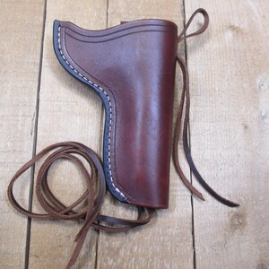 May include: A brown leather holster with white stitching and a dark blue trim. The holster has a loop and a long leather strap. The holster is sitting on a wooden surface.