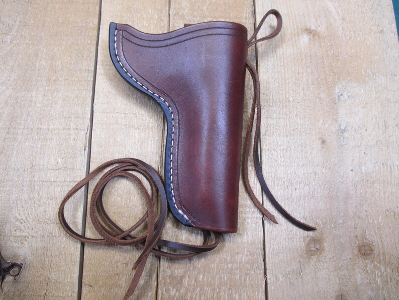 May include: A brown leather holster with white stitching and a dark blue trim. The holster has a loop and a long leather strap. The holster is sitting on a wooden surface.