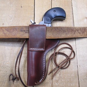 May include: A black handgun is partially visible, resting on a brown leather belt. The belt is threaded through a brown leather holster with white stitching. A leather strap is attached to the holster. The belt is stamped with the words "GENUINE MADE".