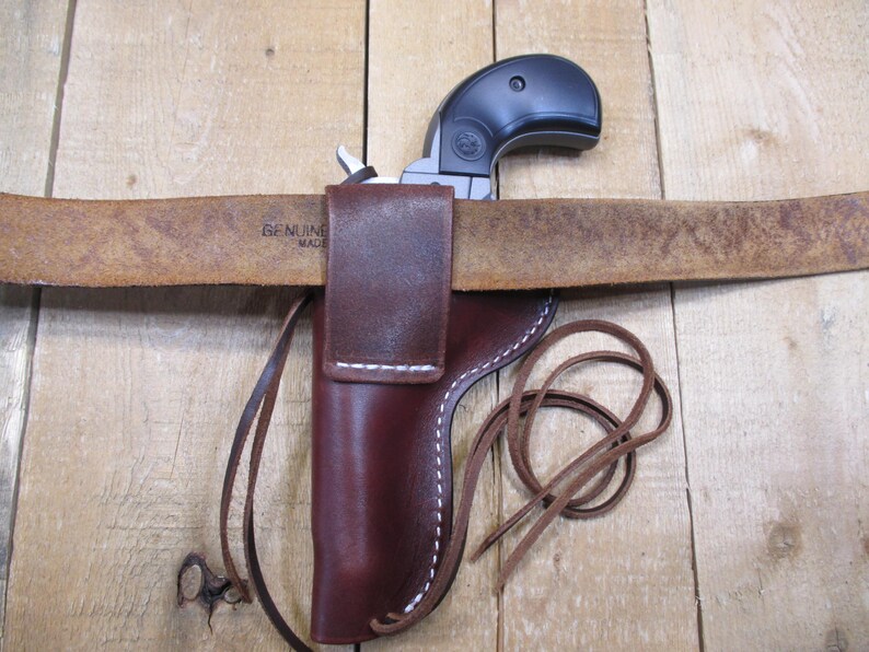 May include: A black handgun is partially visible, resting on a brown leather belt. The belt is threaded through a brown leather holster with white stitching. A leather strap is attached to the holster. The belt is stamped with the words "GENUINE MADE".