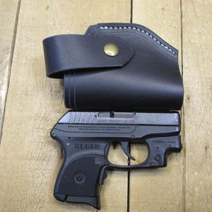 May include: A black leather holster with a brass button and white stitching, paired with a black Ruger handgun. The gun is marked "Ruger Prescott, AZ - USA". The items are displayed on a wooden surface.