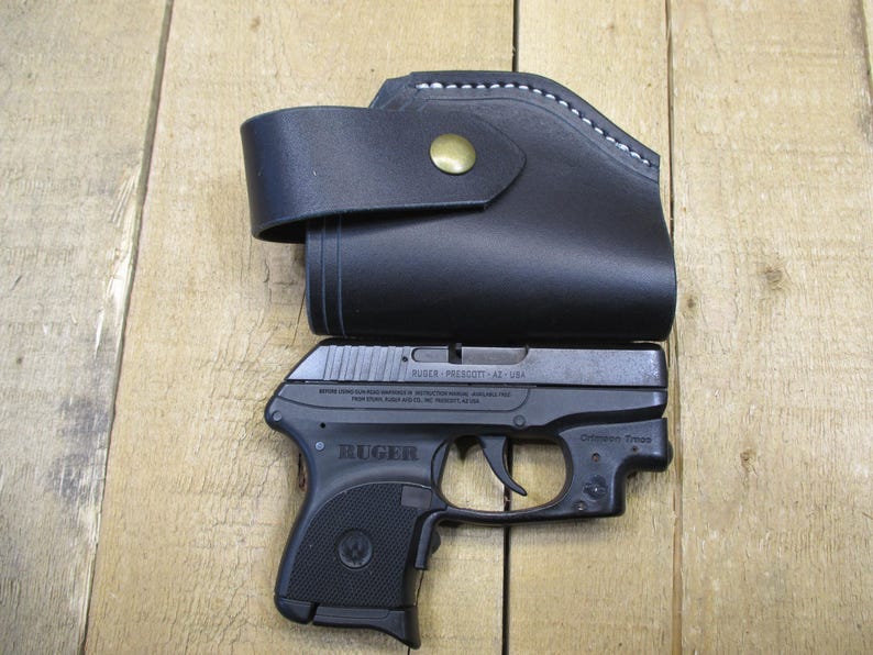 May include: A black leather holster with a brass button and white stitching, paired with a black Ruger handgun. The gun is marked "Ruger Prescott, AZ - USA". The items are displayed on a wooden surface.