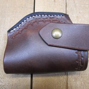 May include: A brown leather holster with a decorative stitched design along the top edge and a strap secured by a brass-colored button. The holster is designed to hold a firearm.