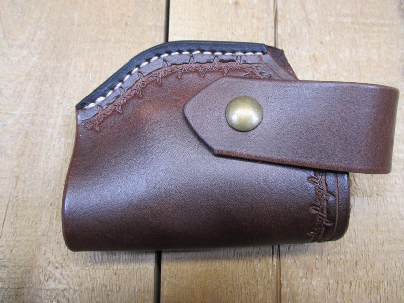 May include: A brown leather holster with a decorative stitched design along the top edge and a strap secured by a brass-colored button. The holster is designed to hold a firearm.
