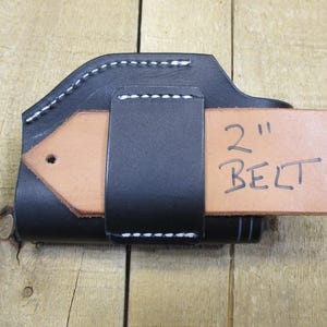 May include: Black leather holster with white stitching, designed to hold a 2-inch belt. The holster has a natural tan leather belt loop. The text "2" and "BELT" are handwritten on the belt loop.