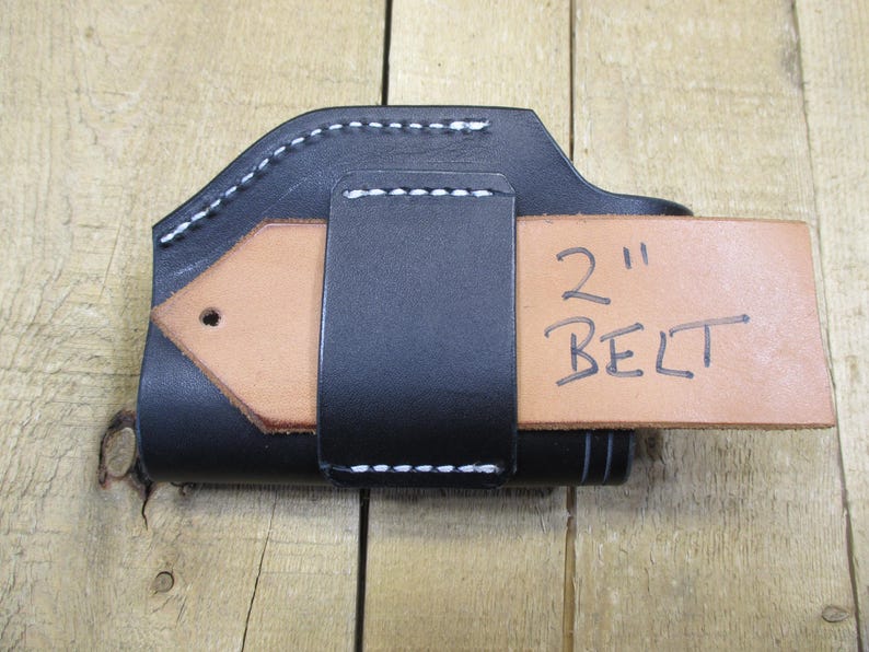 May include: Black leather holster with white stitching, designed to hold a 2-inch belt. The holster has a natural tan leather belt loop. The text "2" and "BELT" are handwritten on the belt loop.