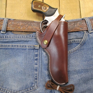 Puede incluir: Una funda de cuero marrón con un revólver, sujeta a unos vaqueros azules. La funda se asegura con un botón de latón y costuras blancas. Se ve un cinturón de cuero marrón con relieve floral.