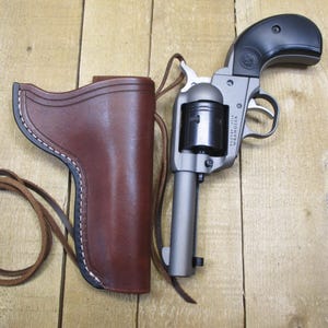 May include: A silver and black revolver with a brown leather holster and a leather strap. The holster is stitched with white thread. The gun is resting on a wooden surface.