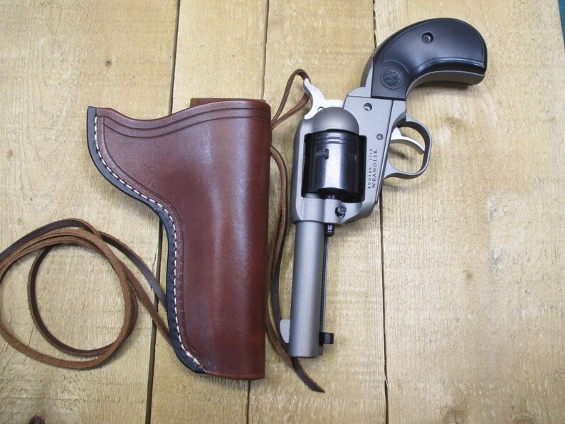 May include: A silver and black revolver with a brown leather holster and a leather strap. The holster is stitched with white thread. The gun is resting on a wooden surface.