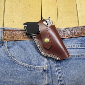 May include: A brown leather holster with a snap closure, holding a silver handgun. The holster is worn on a pair of blue jeans with a brown leather belt.