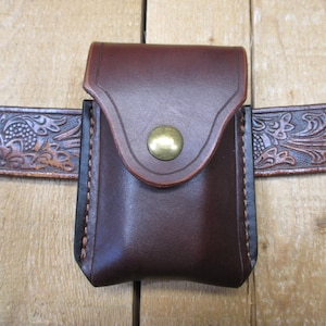 Op de afbeelding: Een donkerbruin leren etui met een klep en een messing knoopsluiting, bevestigd aan een bruine, gesneden leren riem. Het etui is gestikt met lichtgekleurd garen en heeft een rechthoekige vorm. De riem heeft een bloemenpatroon.