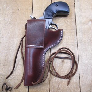 May include: A black and silver handgun rests in a dark brown leather holster. The holster has white stitching and a flap closure. A leather tie is coiled next to the holster. The background is a light brown wooden surface.