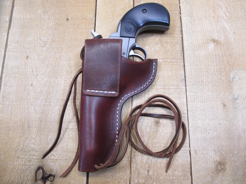 May include: A black and silver handgun rests in a dark brown leather holster. The holster has white stitching and a flap closure. A leather tie is coiled next to the holster. The background is a light brown wooden surface.