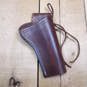 May include: A brown leather holster with white stitching along the edges. The holster has a loop at the top and a leather tie. The leather has a smooth, polished finish.