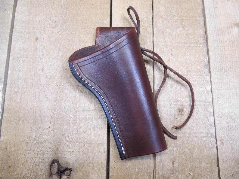 May include: A brown leather holster with white stitching along the edges. The holster has a loop at the top and a leather tie. The leather has a smooth, polished finish.